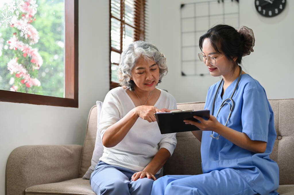 Healthcare professional in blue scrubs reviewing a clipboard with an older woman at home.