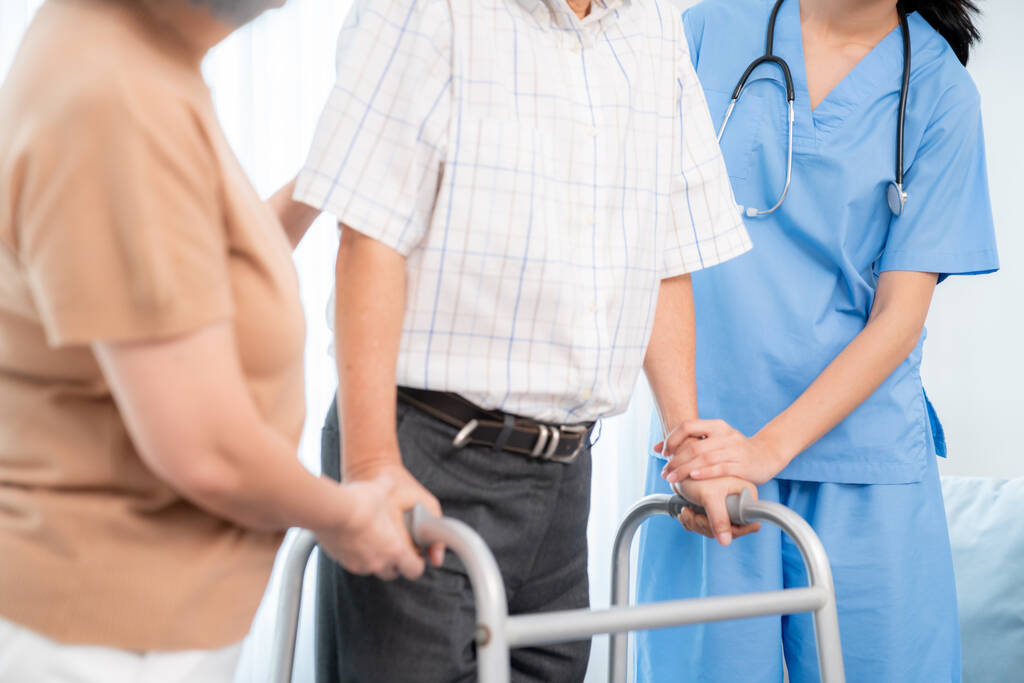 Elderly man using a walker assisted by a caregiver in scrubs and another woman.